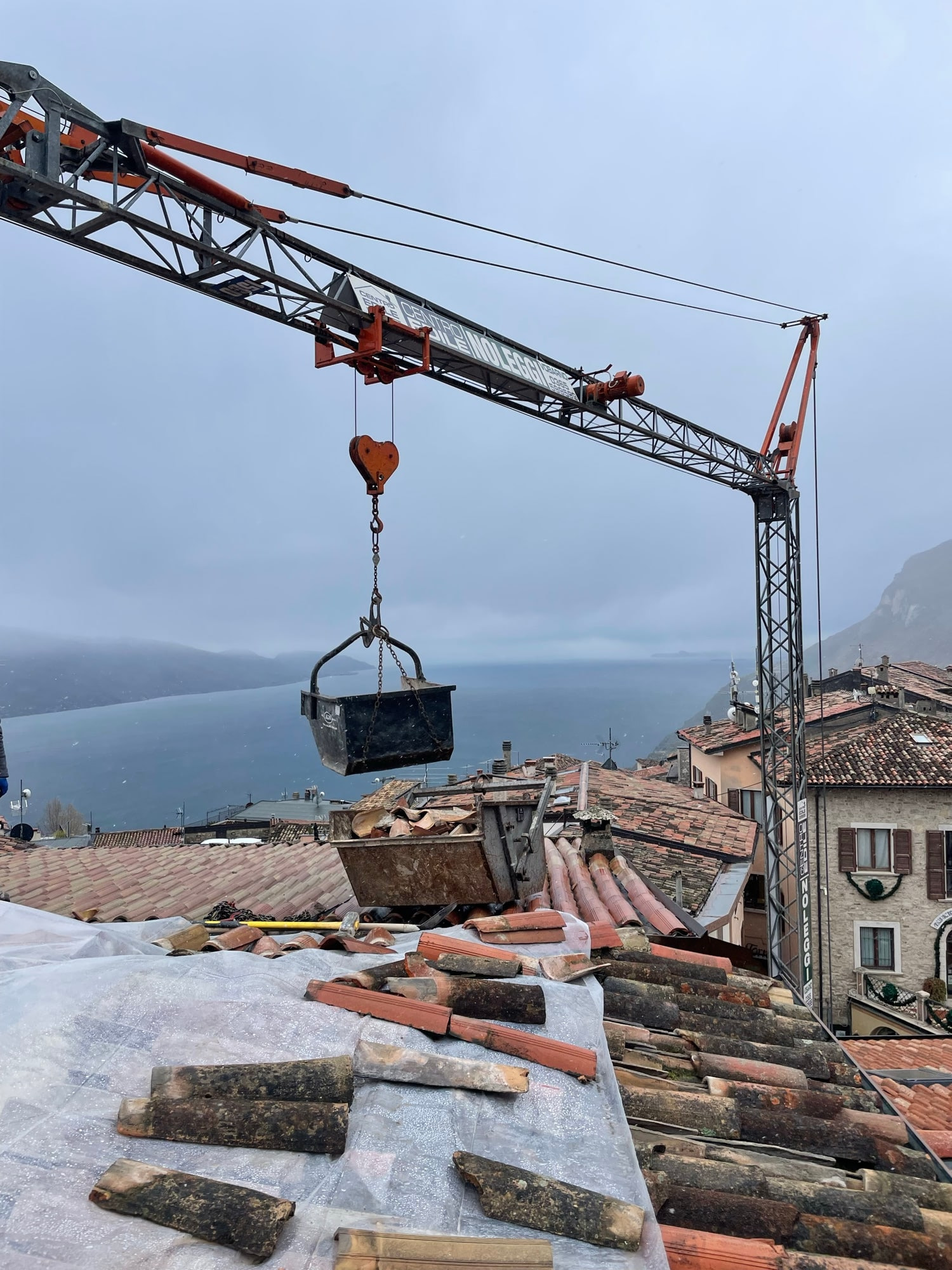Smontaggio del tetto: la gru cala i coppi vecchi in un cassone, sullo sfondo il Lago di Garda