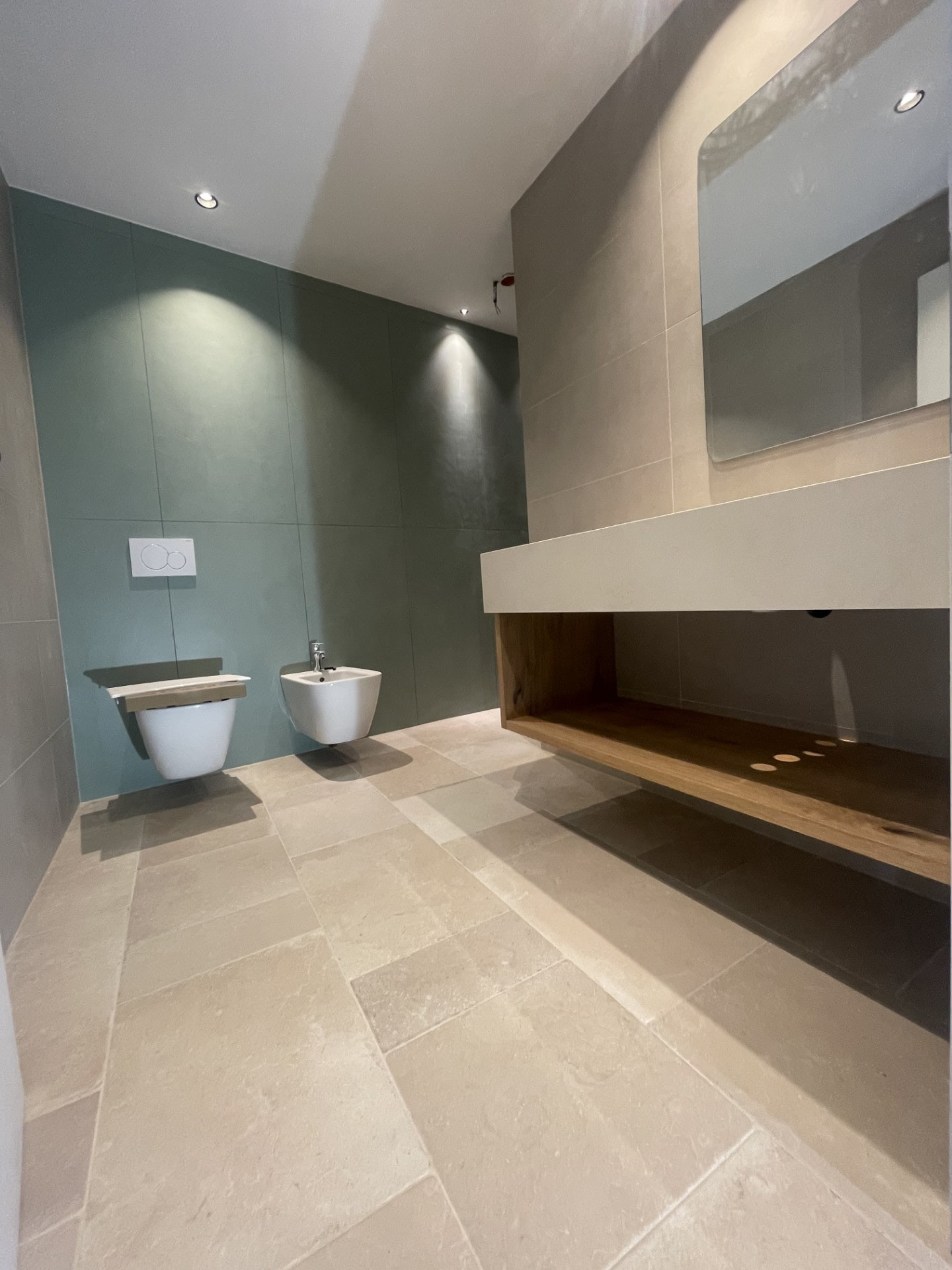 Bagno con parete verde in grandi lastre, sanitari sospesi, lavabo su mobile in legno e pavimento in pietra