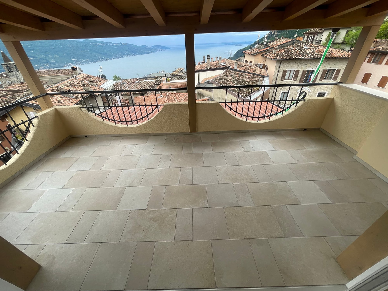 Terrazza coperta con travi in legno, pavimento in pietra e ringhiera in ferro, con vista sui tetti e sul Lago di Garda