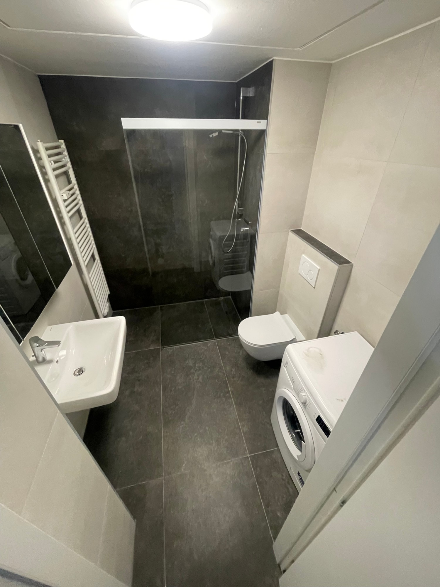 Bagno finito visto dalla porta: piastrelle scure di grande formato, doccia, WC sospeso e lavatrice